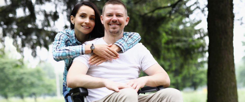 Smiling couple outdoors, the man in a wheelchair and his partner hugging him