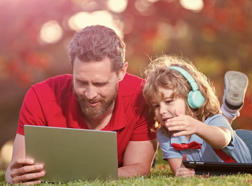 Banner of father on son school boy with laptop study online lying on grass, father and son use modern communication technology in park. family blog.